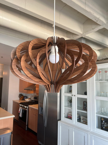 May include: A wooden pendant light fixture with a unique, geometric design. The light is made of thin, curved pieces of wood that are arranged in a circular pattern. The light is hanging from a white cord and has a white light bulb.
